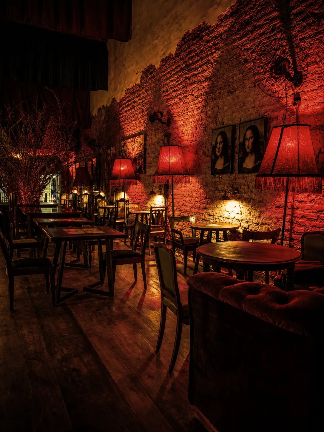 Parede de tijolo colonial do BICA BAR com as releituras da Mona Lisa e luminária de cúpula vermelha, sob iluminação cênica baixa